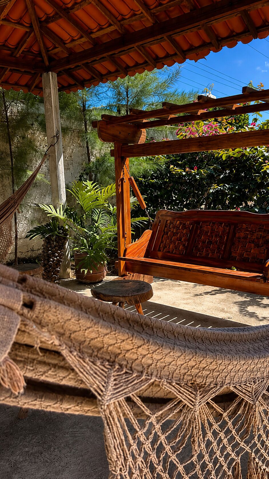 A hammock, wooden bench, and potted plants under a wooden pergola.