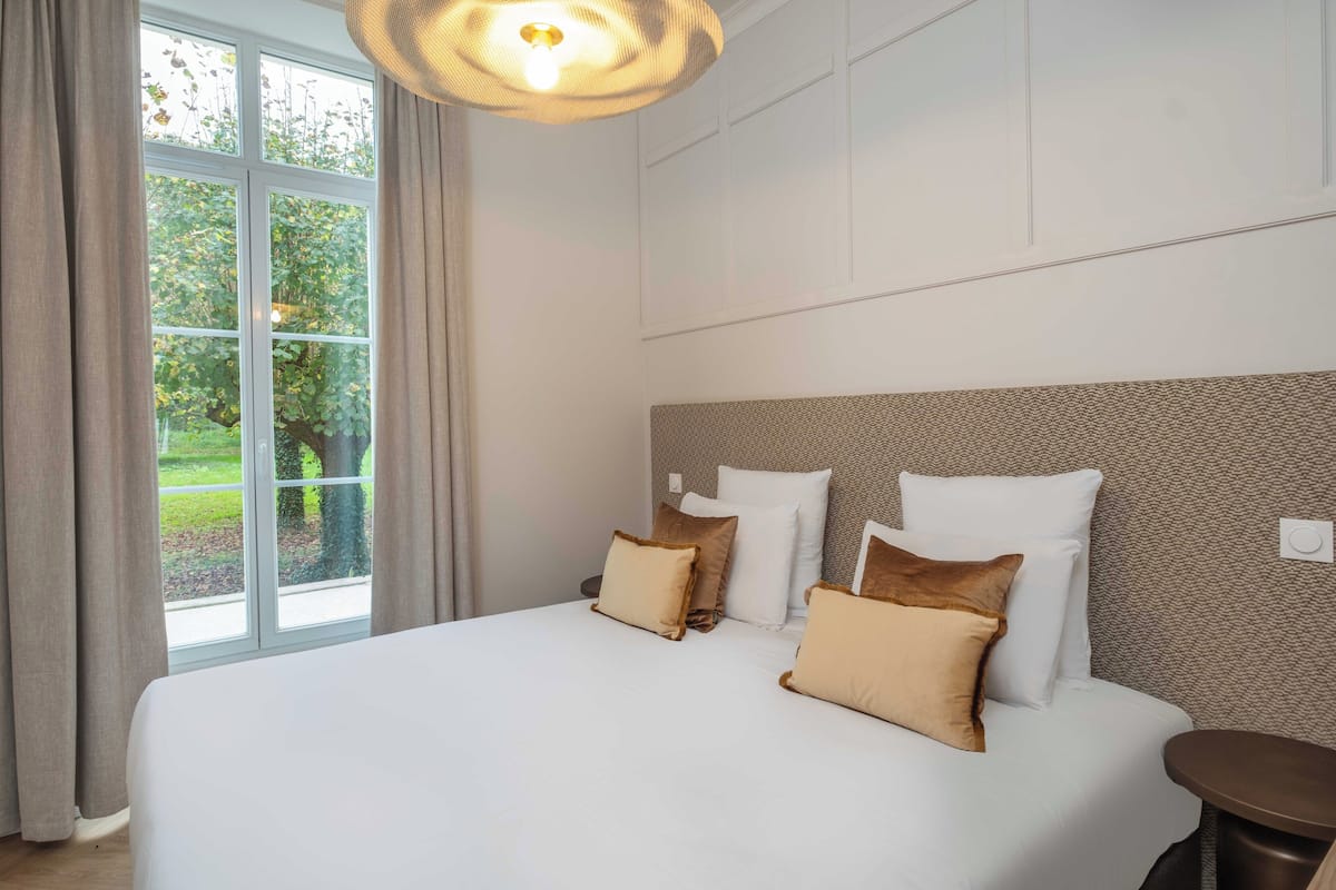 A neatly made bed with white and beige pillows, a round bedside table, and a window with curtains.
