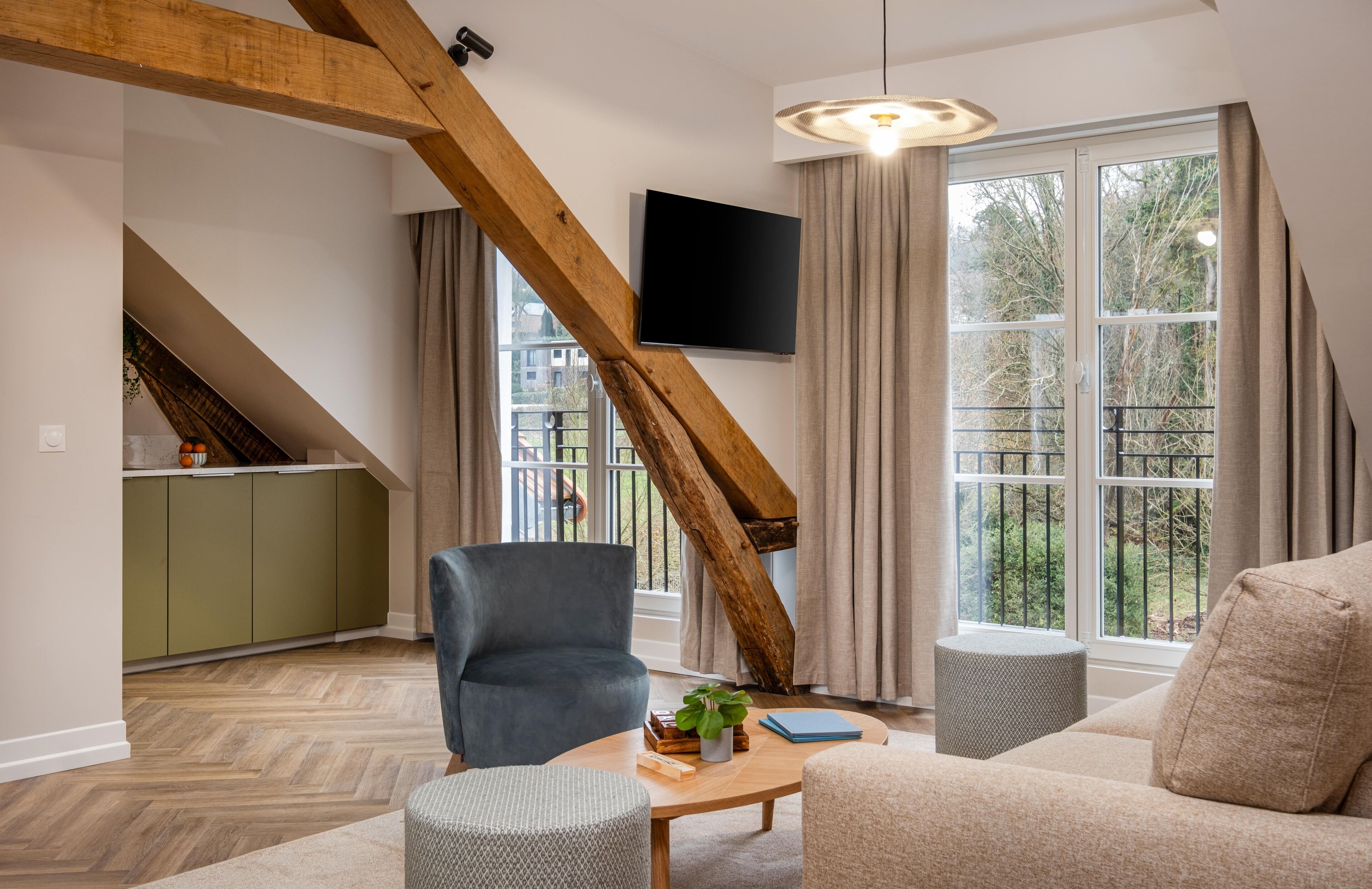 Ein modernes Wohnzimmer mit Holzbalkendecke, einem Flachbildfernseher, einem Sofa, einem Sessel und einem Couchtisch mit Büchern und einer Pflanze.