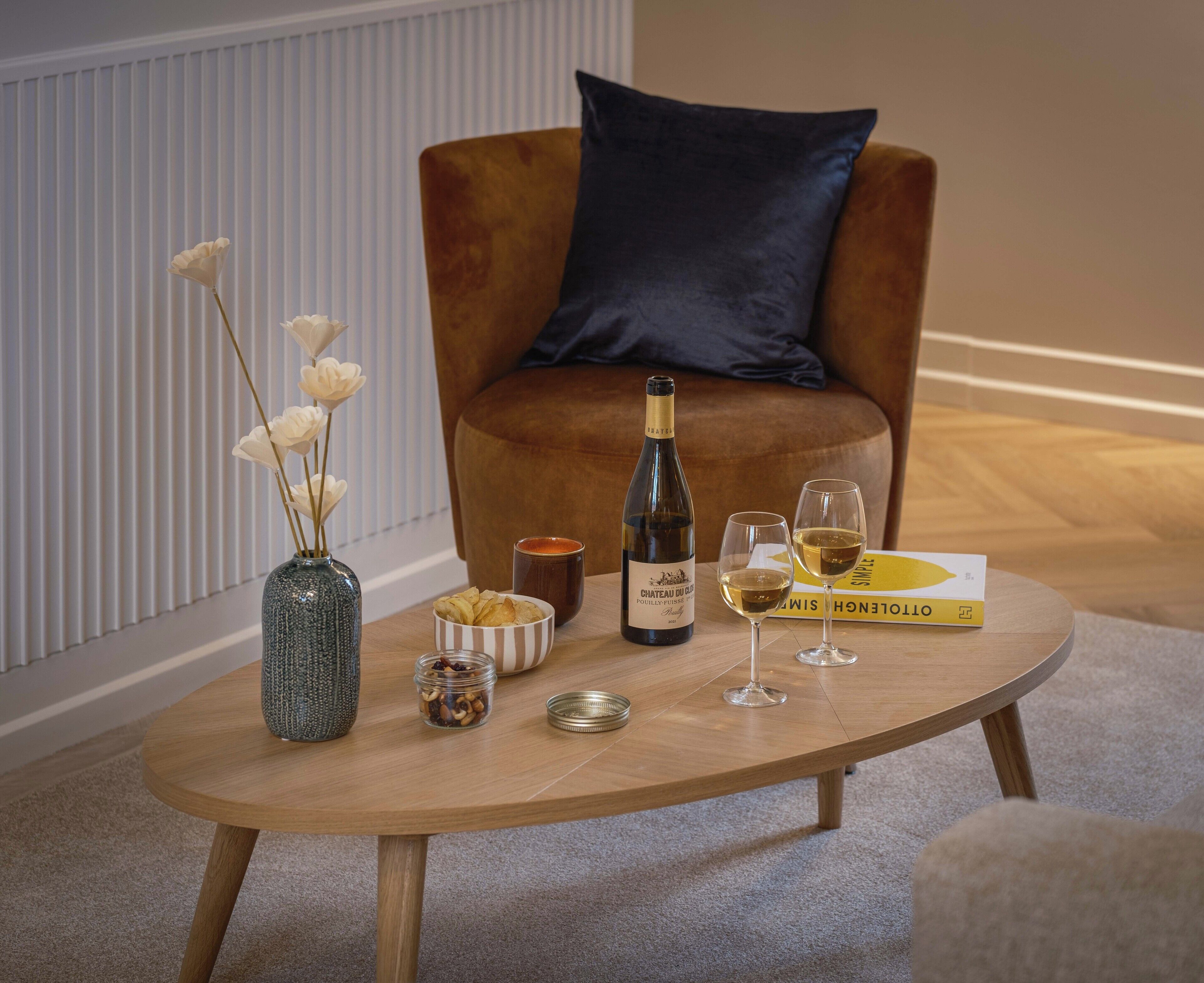 Een ronde houten tafel met een fles wijn, twee wijnglazen, een schaal met snacks, een potje noten en een vaas met bloemen.