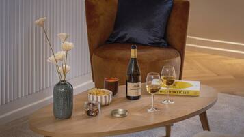 Een ronde houten tafel met een fles wijn, twee wijnglazen, een schaal met snacks, een potje noten en een vaas met bloemen.