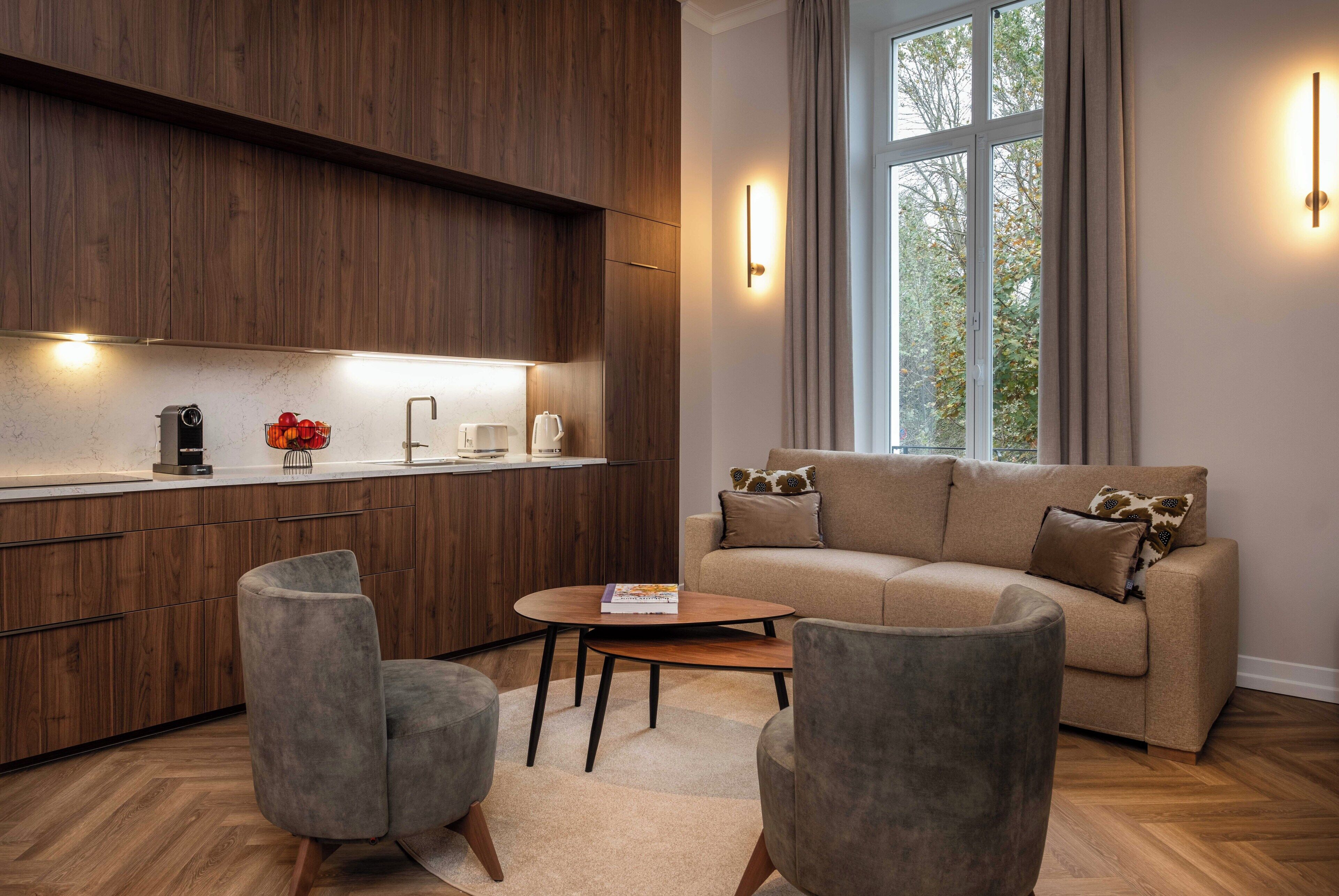 A modern living room with a sofa, two armchairs, a coffee table, and a kitchen area with wooden cabinets and a window.