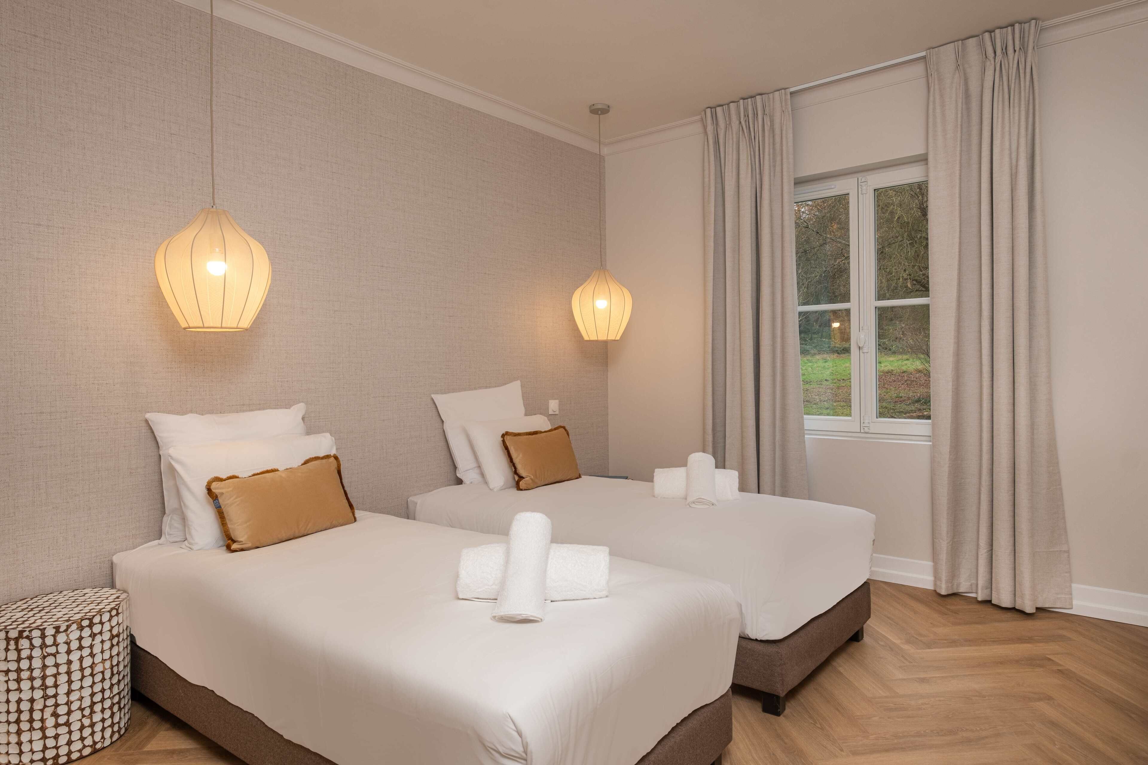 A hotel room with two beds, a window with curtains, and a hanging light fixture.