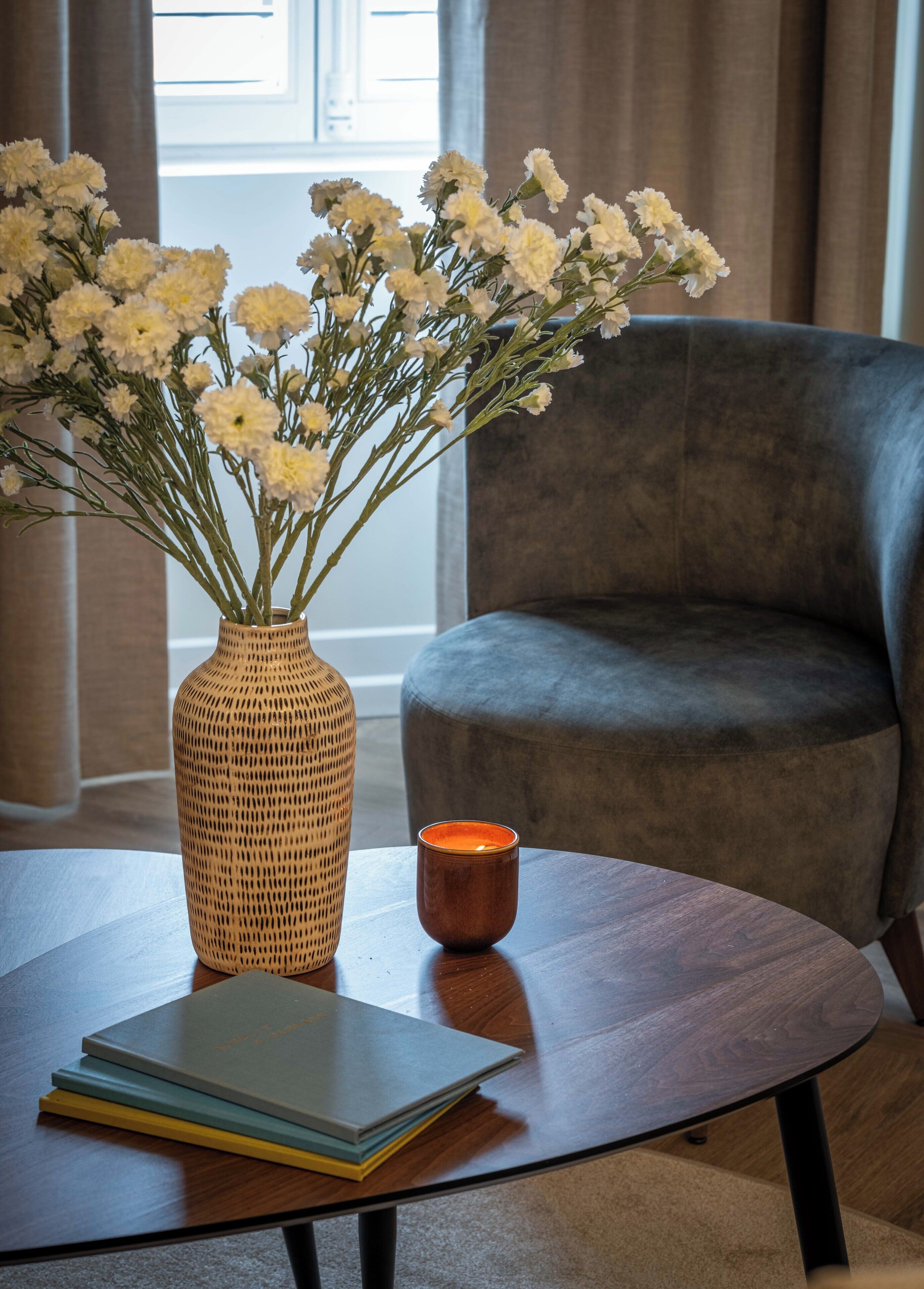 A room with a dark grey armchair, a wooden table with a vase of white flowers, and a lit candle.