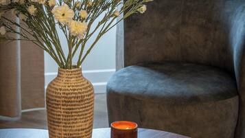 A room with a dark grey armchair, a wooden table with a vase of white flowers, and a lit candle.
