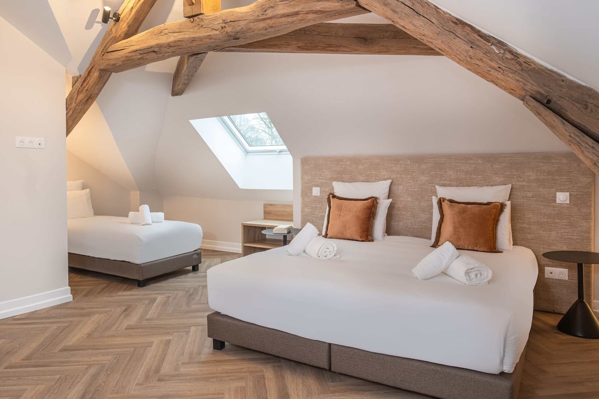 A bedroom with a large bed, a smaller bed, wooden beams, a skylight, and a herringbone floor pattern.