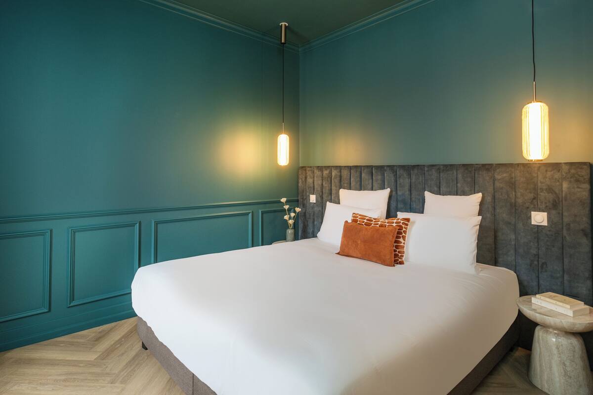 A neatly made bed with white linens and a brown accent pillow, a wooden headboard, teal walls, and two pendant lights.