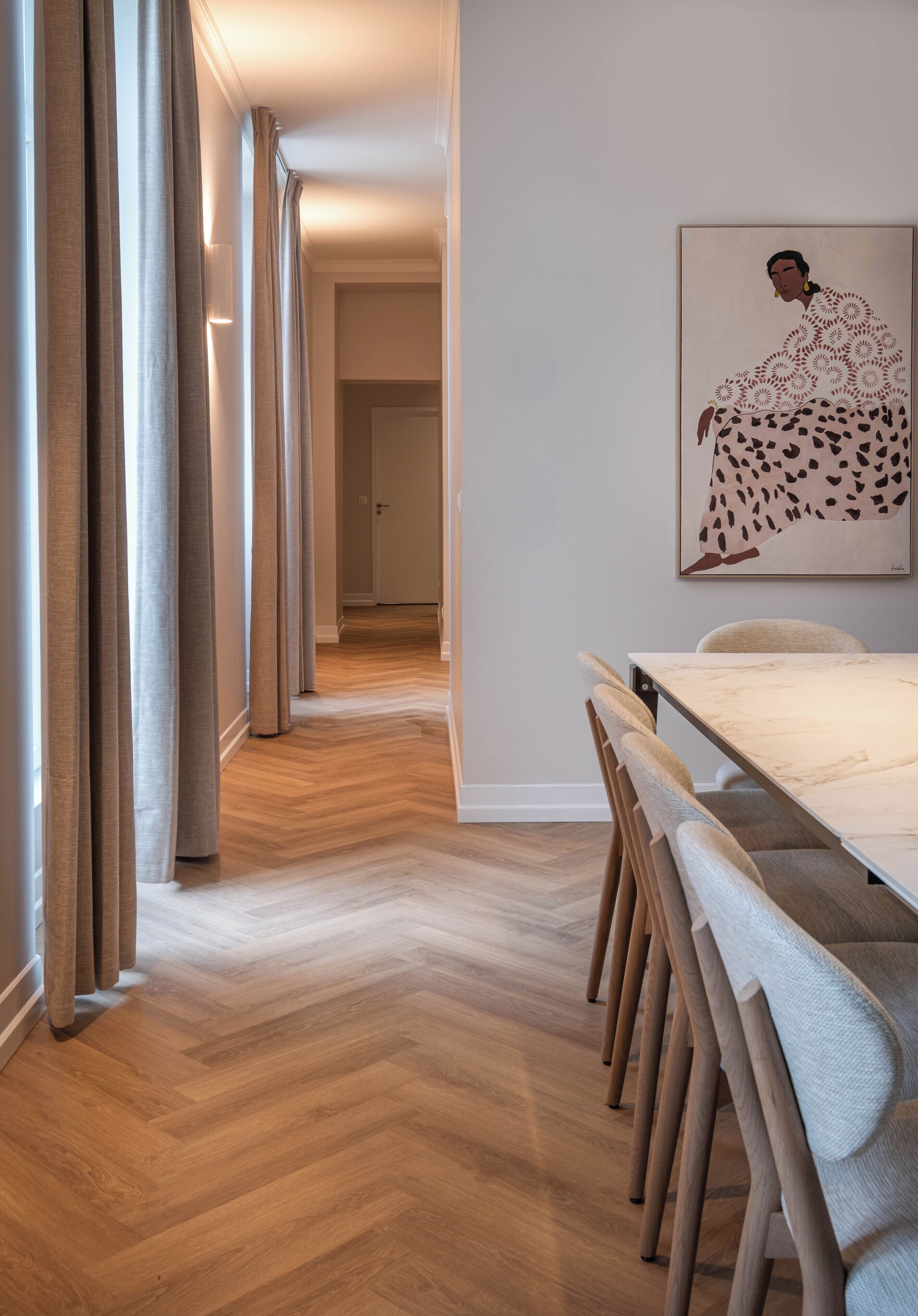 A modern interior with a wooden herringbone floor, a dining area with a marble table and upholstered chairs, and a large abstract painting on the wall.