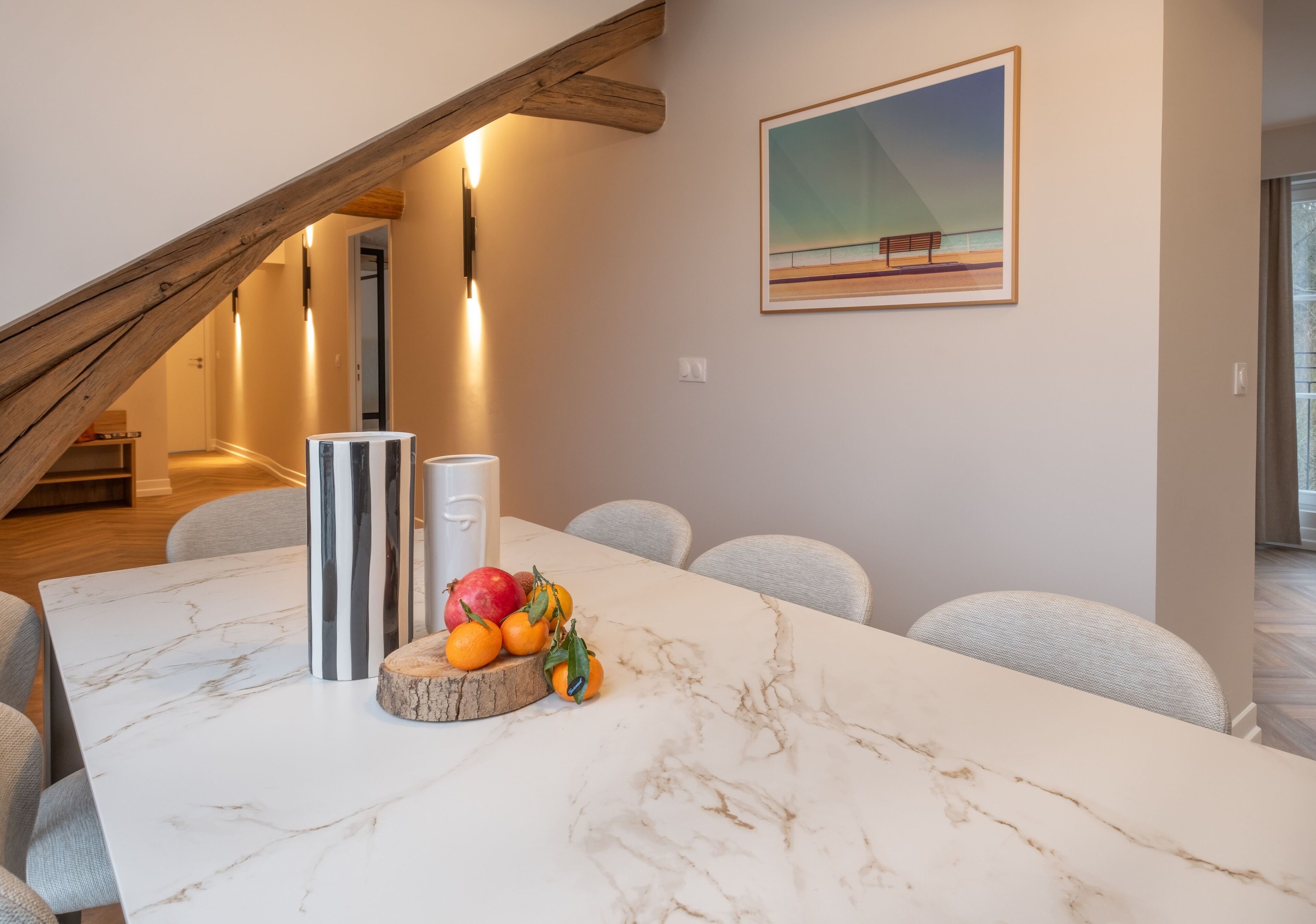 A modern dining area with a marble table, fruit centerpiece, and a painting on the wall.