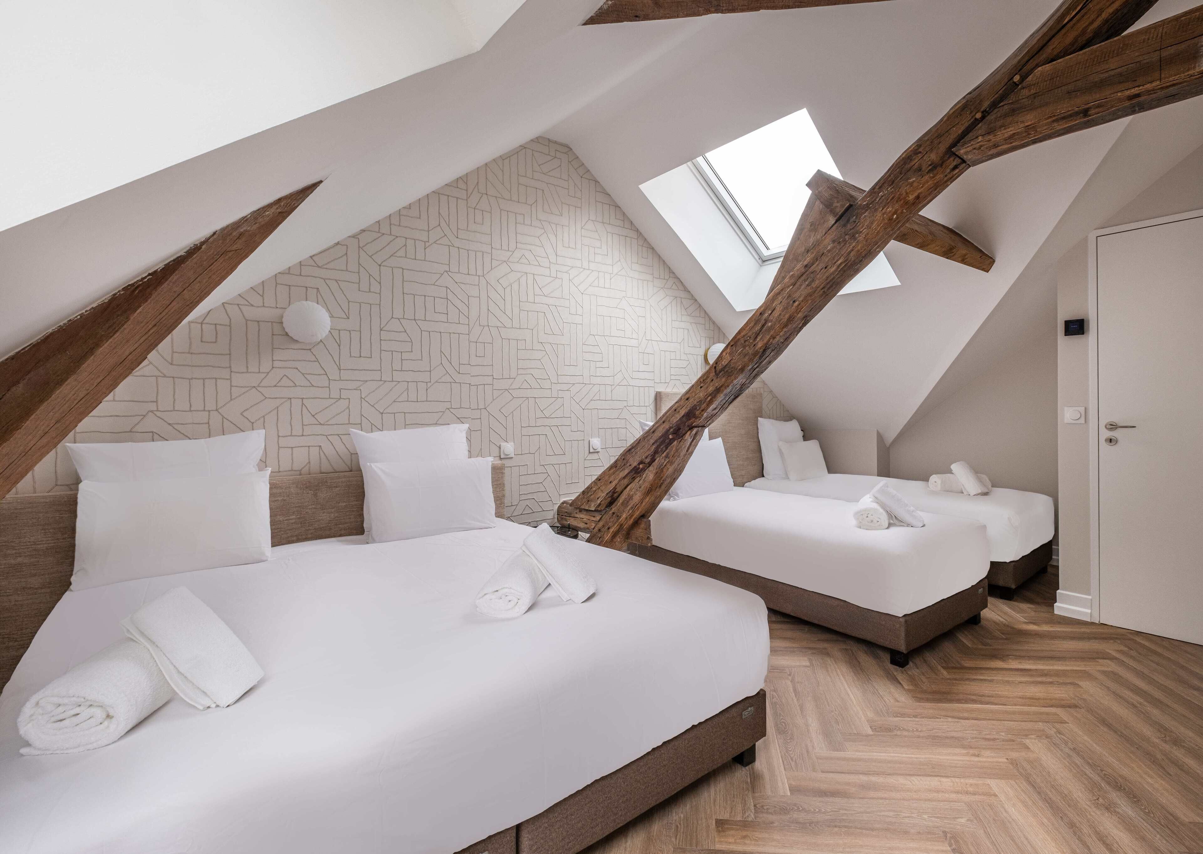 A modern loft bedroom with two beds, wooden beams, a geometric patterned wall, and a skylight.