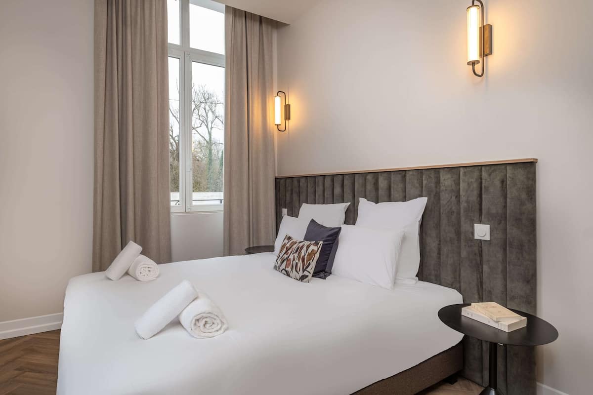 A neatly made bed with pillows and rolled towels, a bedside table with a book, and a window with curtains.