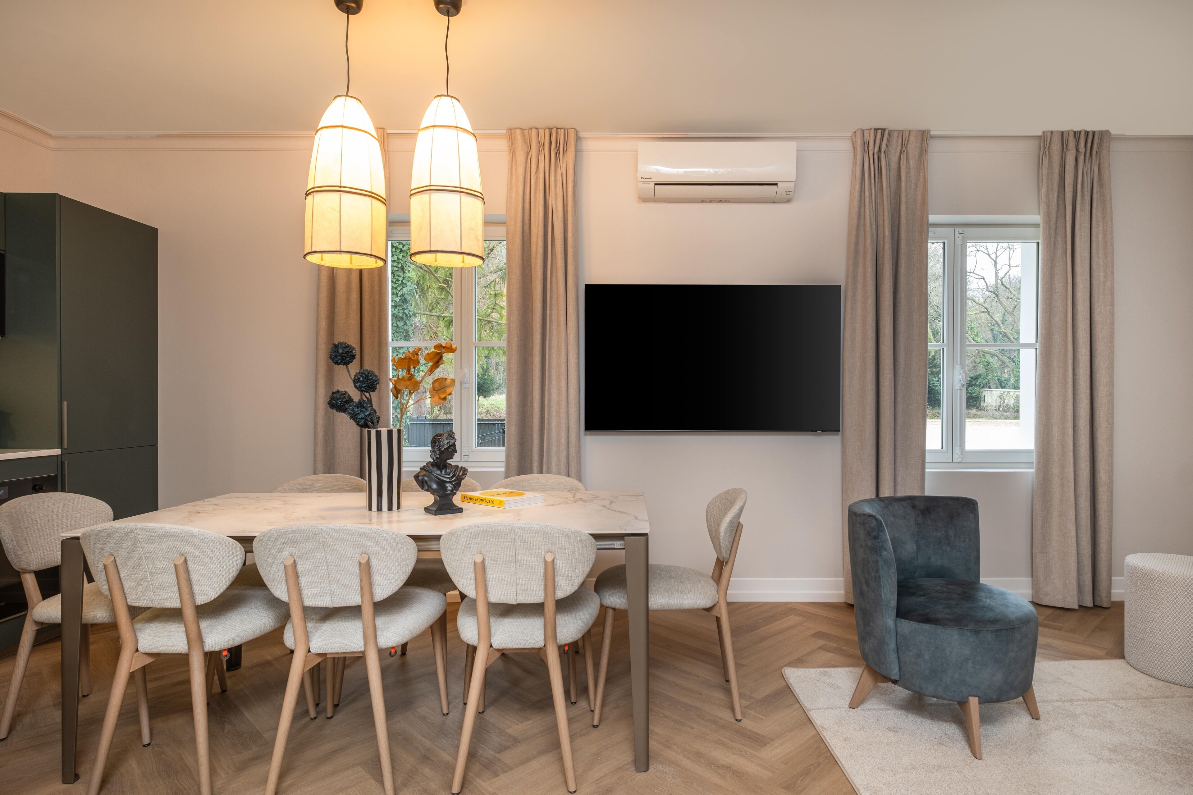 Un comedor moderno con una mesa y sillas de madera, un televisor de pantalla plana montado en la pared y un ventanal con cortinas.