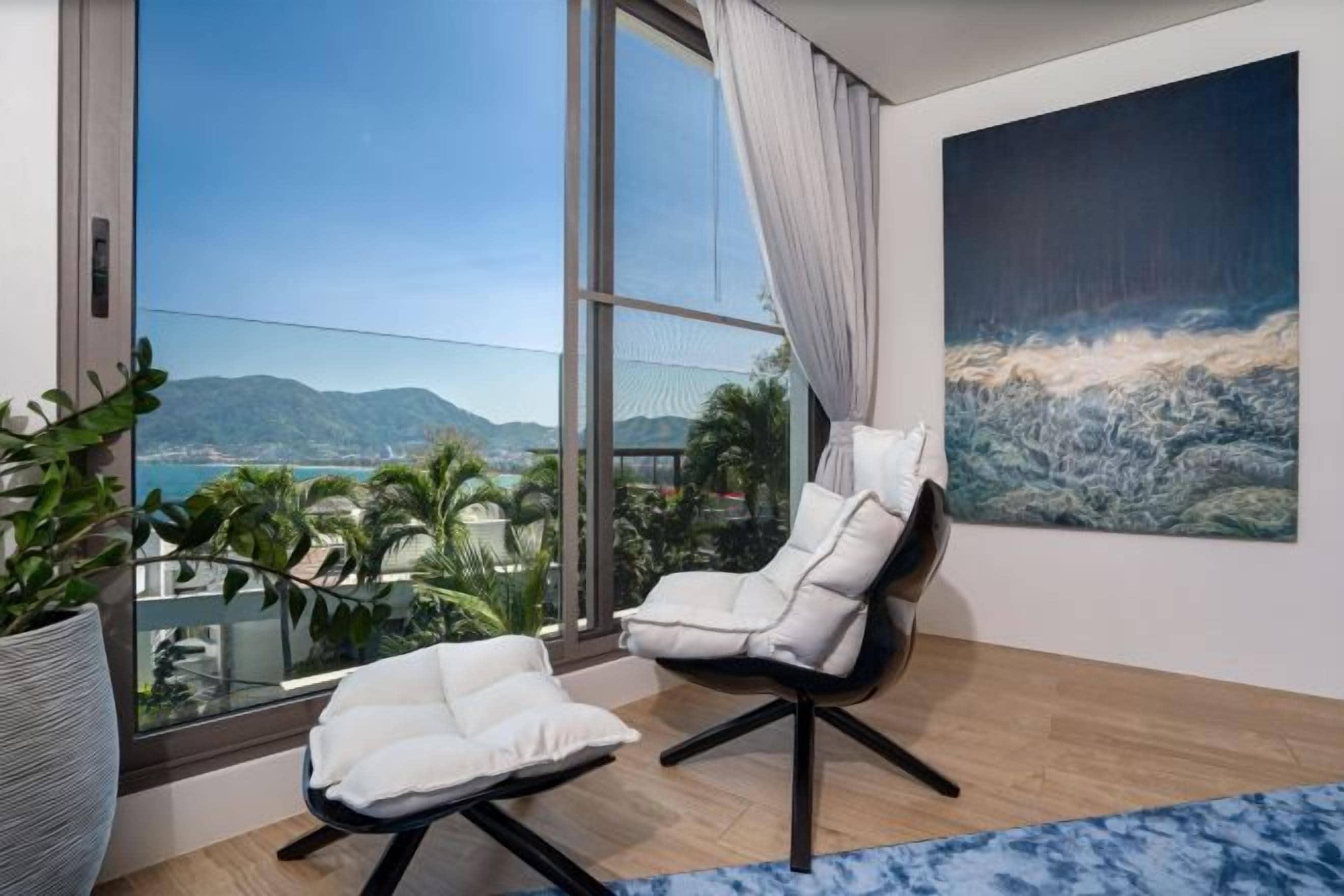 A modern living room with a large window, two white armchairs, a glass table, and a painting of a coastal scene.