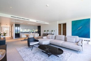 A modern living room with a sectional sofa, a black coffee table, and a dining area with a kitchen in the background. - New 2Br Condo Bluepoint 8-17 Sea Views (Phuket)