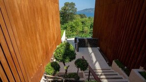 Vista a través de un corredor de madera con un banco de piedra y árboles pequeños, que conduce a un área exterior panorámica con vistas al mar y colinas.
