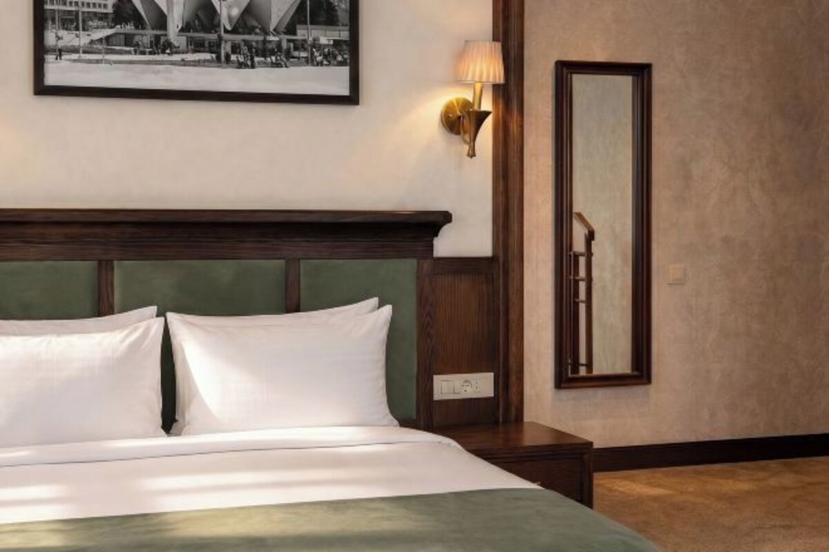 A hotel room with a bed, a nightstand, a wall-mounted lamp, a framed picture, and a mirror.