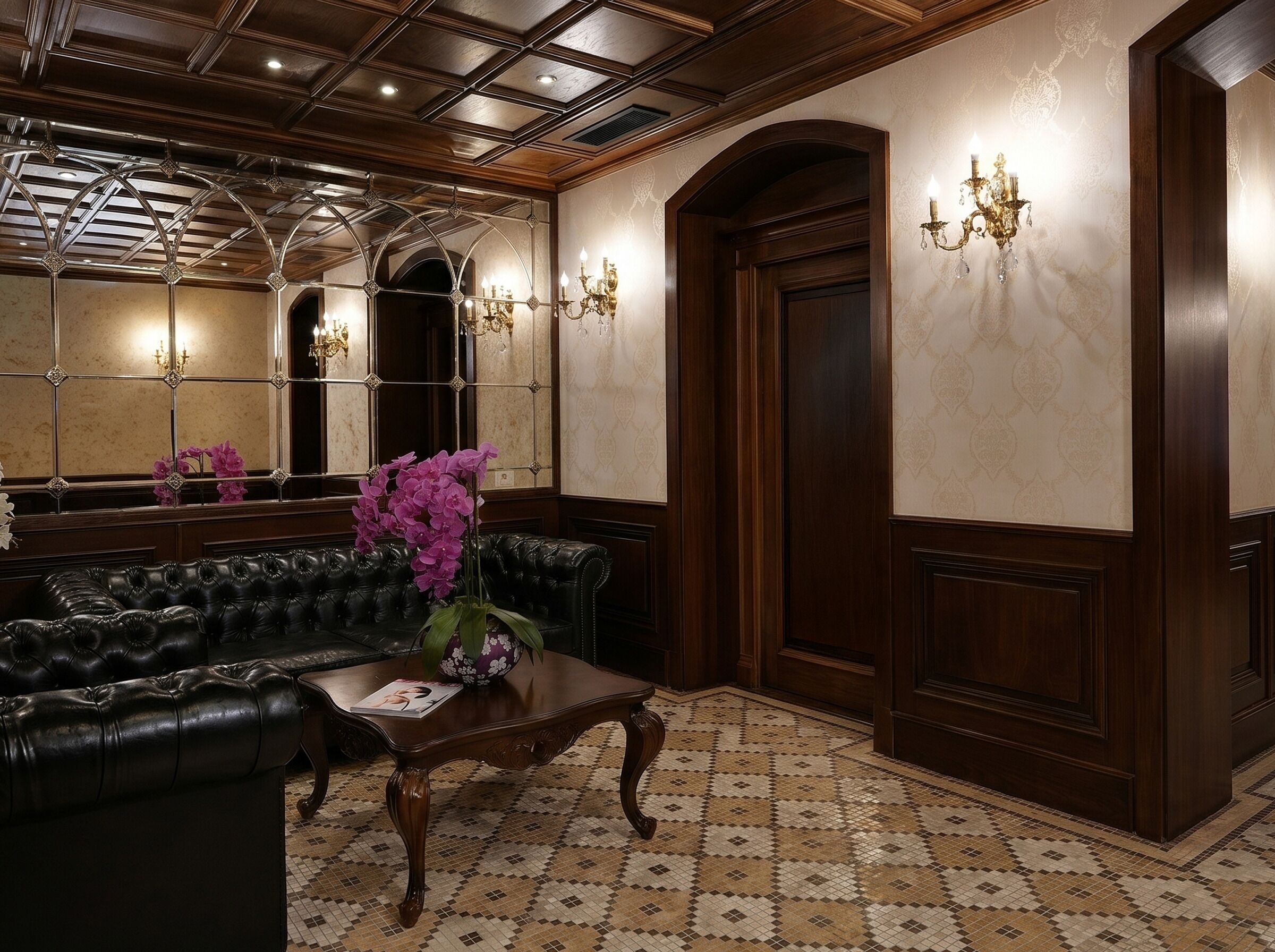 Un vestíbulo con un sofá de cuero negro, una mesa de centro de madera con un jarrón de flores, un suelo de baldosas con diseño y un espejo grande con apliques.