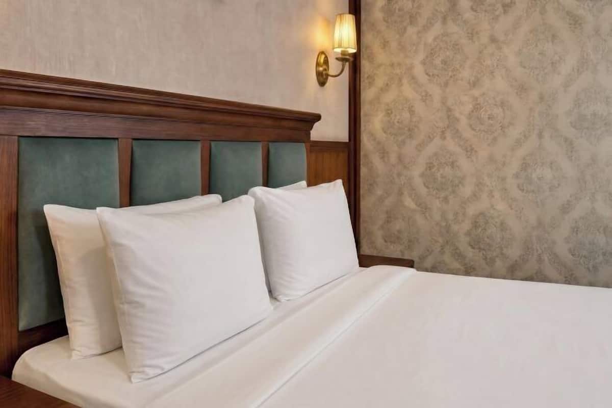 A neatly made bed with white linens and pillows, a wooden headboard, and a wall-mounted light fixture.