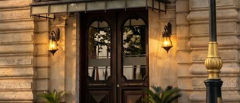 L’entrée d’un hôtel, avec une façade en bois sombre, deux plantes en pot et deux lampes fixées au mur.