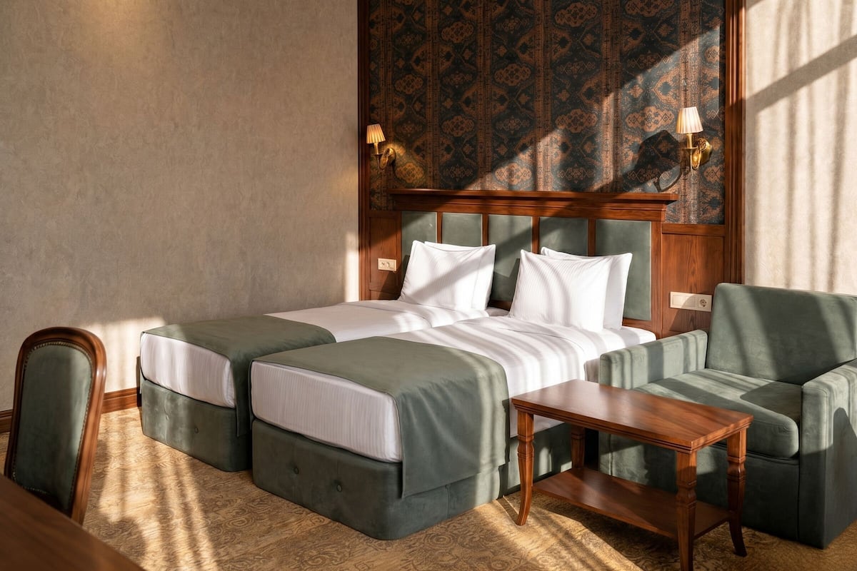 A hotel room with two beds, a wooden headboard, a green sofa, a wooden table, and patterned wallpaper.