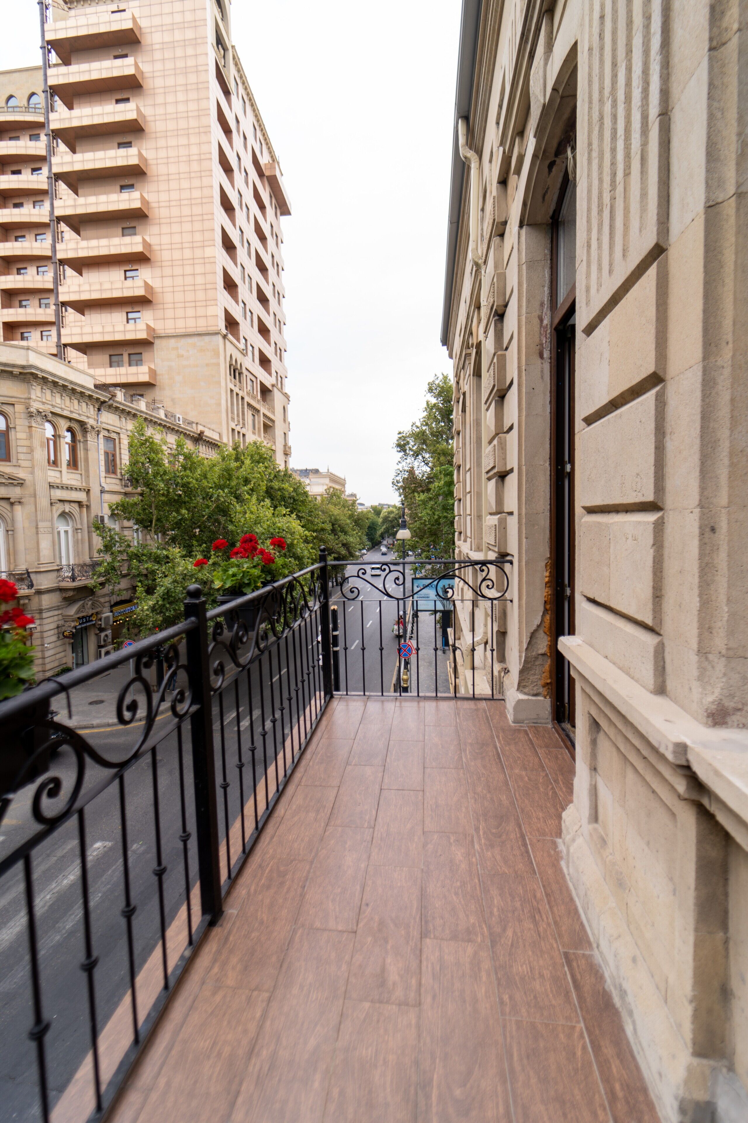 A narrow balcony with a black wrought iron railing, a tiled floor, and a view of a city street with buildings and trees.