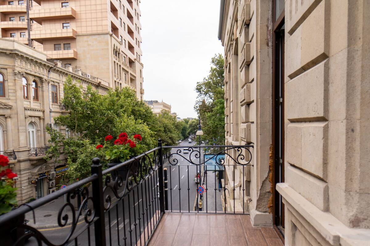 A narrow balcony with a black wrought iron railing, a tiled floor, and a view of a city street with buildings and trees.