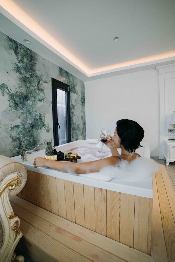 Uma pessoa relaxando em uma banheira com uma bebida, em um banheiro moderno com piso de madeira e uma janela grande.