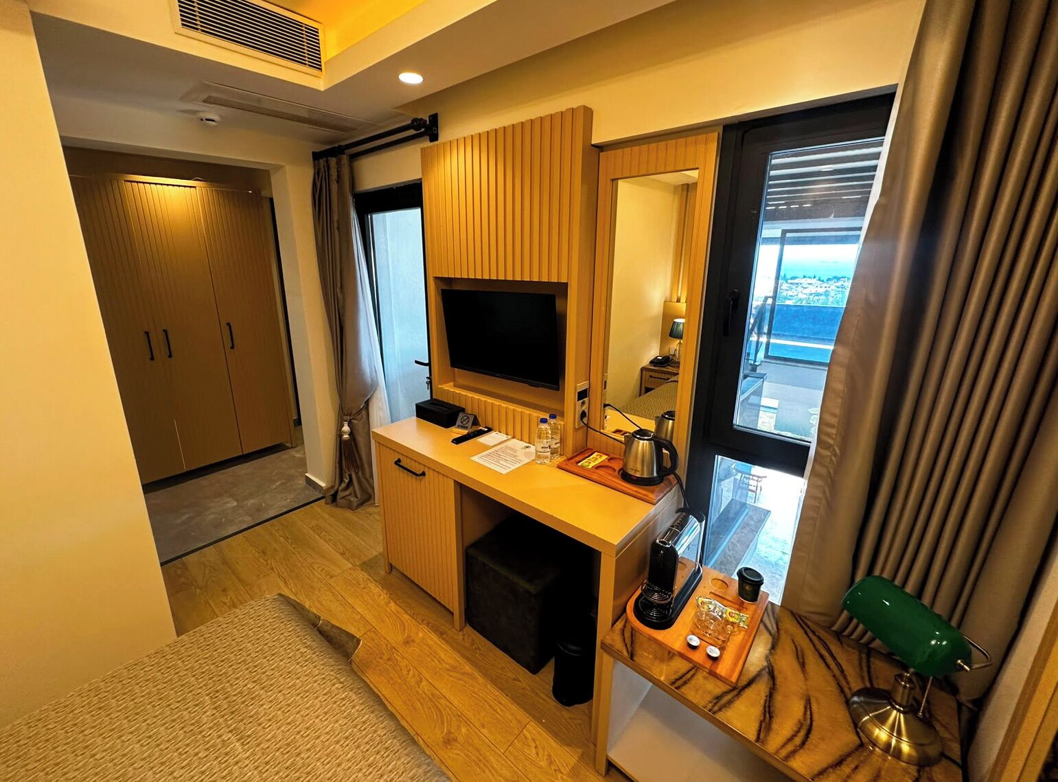 Habitación de hotel con escritorio de madera, televisor, espejo, ventana con cortinas y vista al mar.