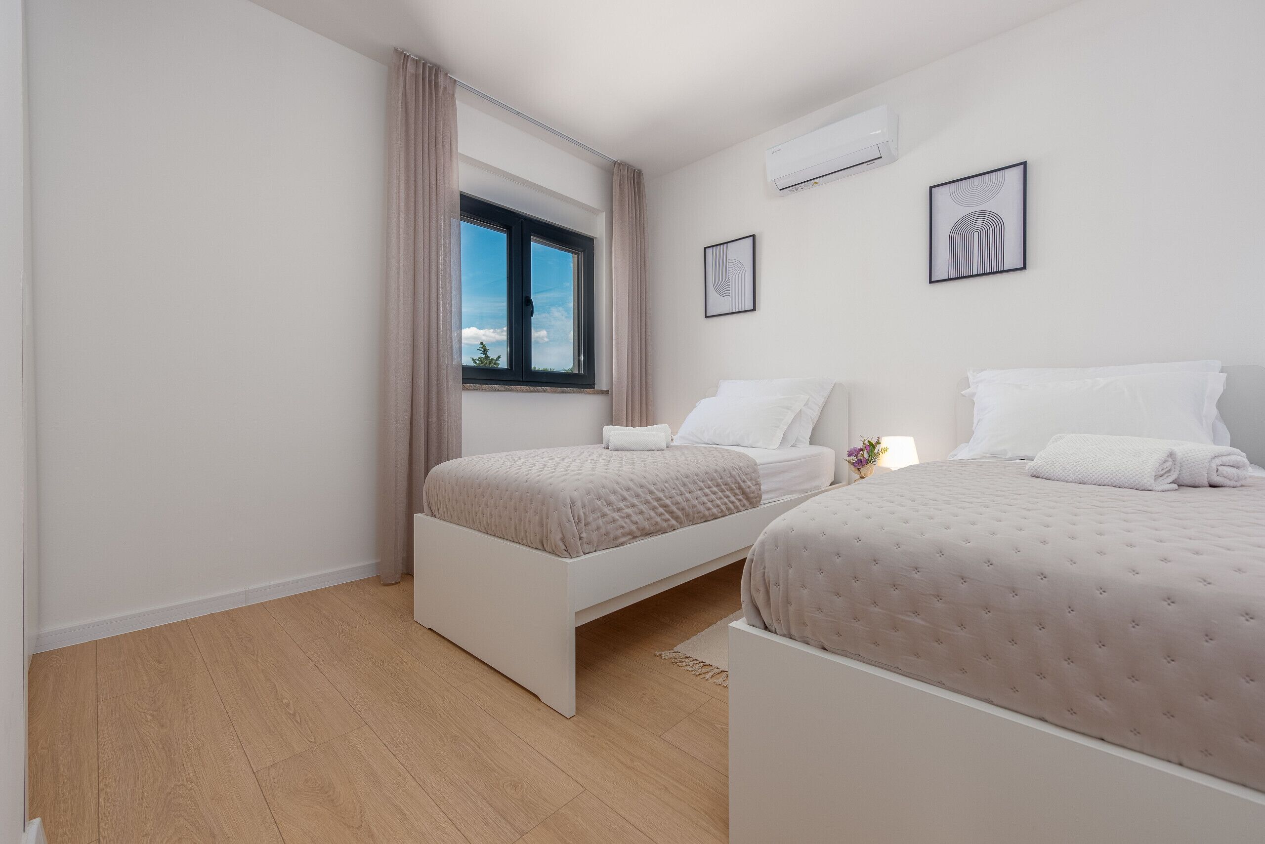 A modern bedroom with two beds, a window with curtains, and a wall-mounted air conditioner.