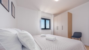 Una cama bien hecha con edredón blanco y almohadas, una ventana con cortinas, un armario de madera y una silla.