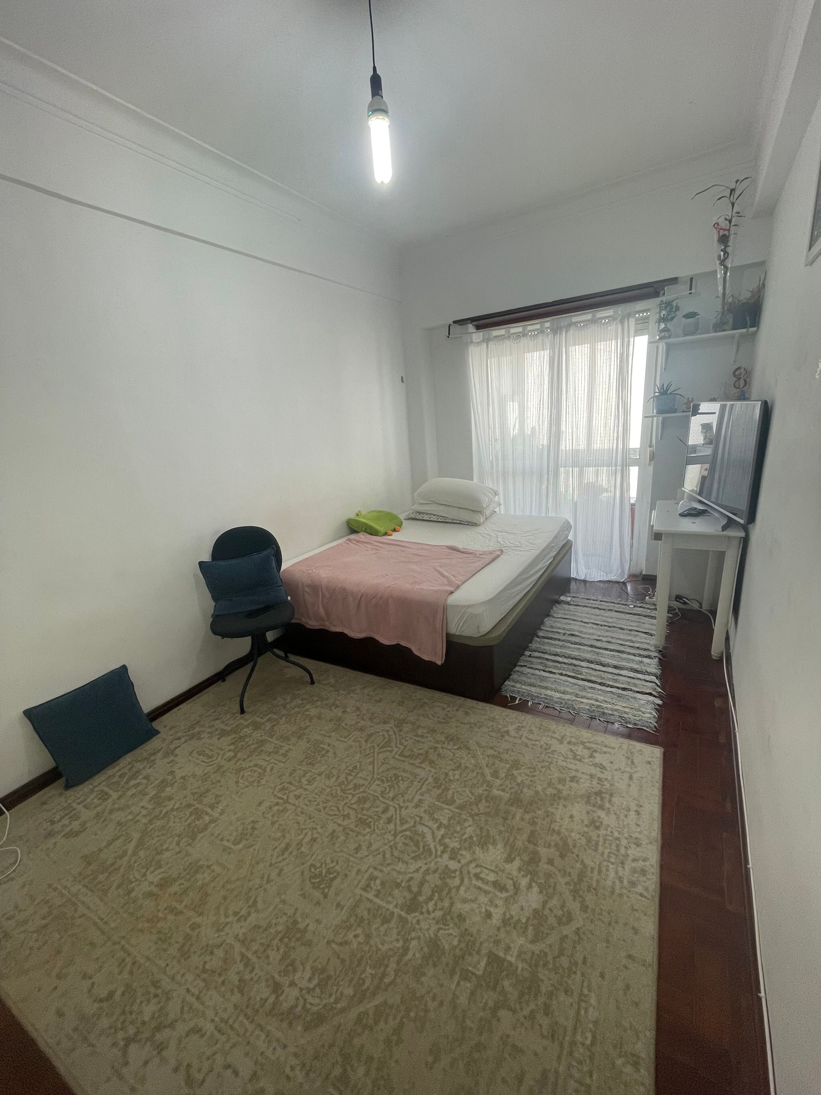 A bedroom with a bed, a desk, a chair, a TV, and a hanging light fixture.
