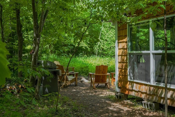 Una cabaña rústica con un ventanal grande, rodeada de un bosque frondoso. Hay dos sillas de madera y una mesita afuera.