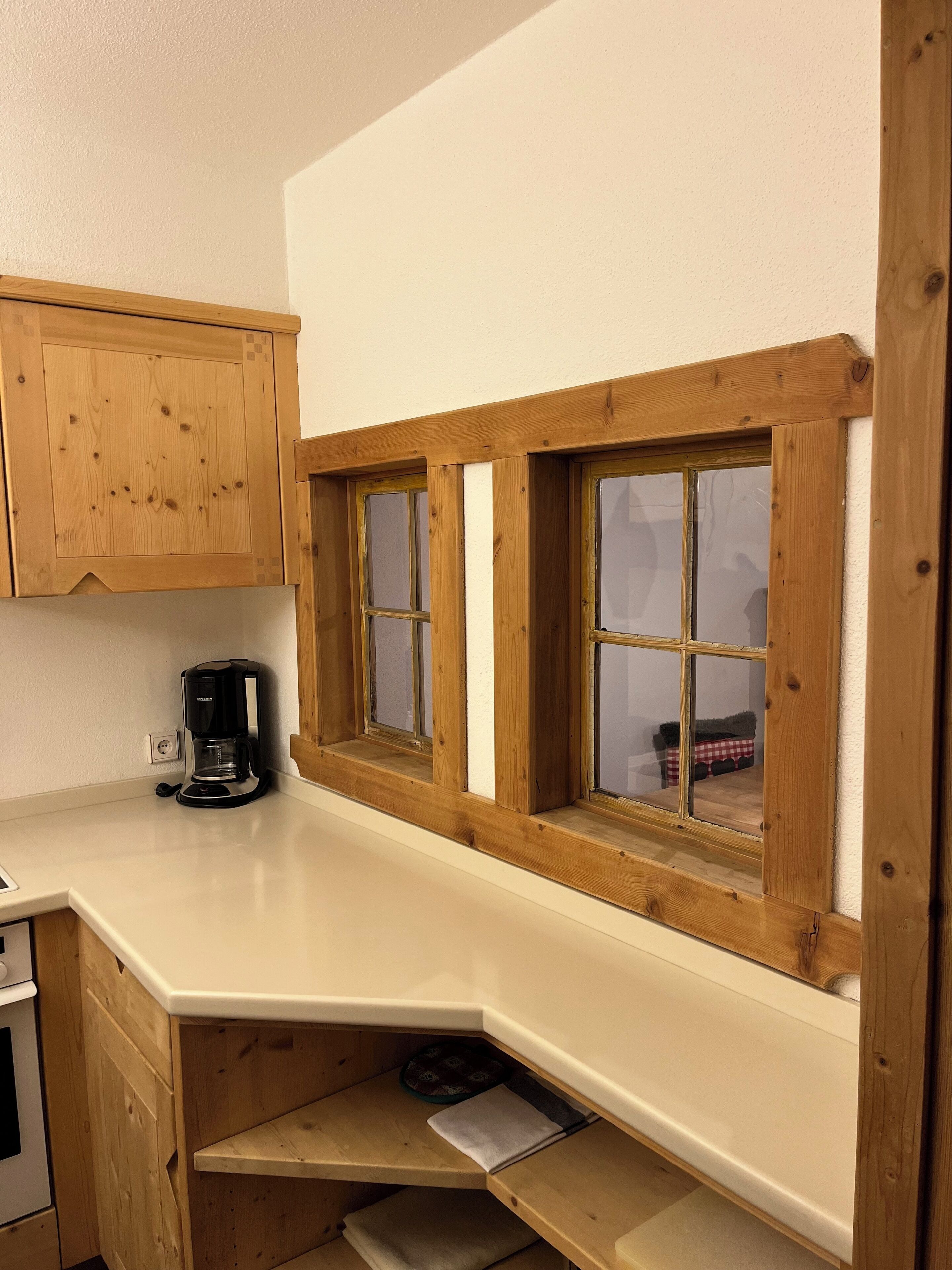 Une cuisine avec des meubles en bois, une machine à café et une fenêtre donnant sur une table recouverte d’une nappe à carreaux rouges et blancs.