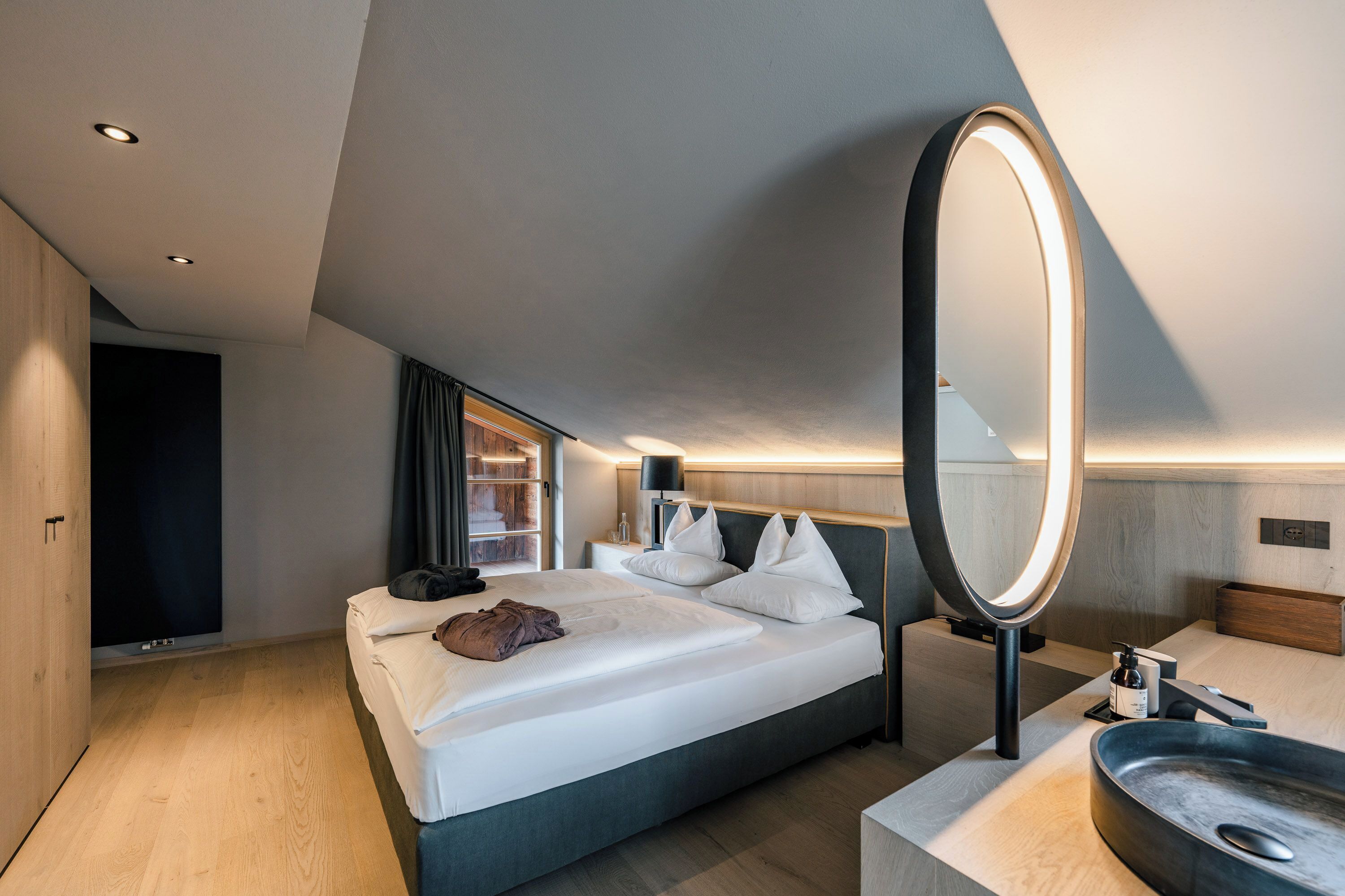Una habitación de hotel moderna con una cama grande, mesita de noche y baño con lavamanos y espejo.