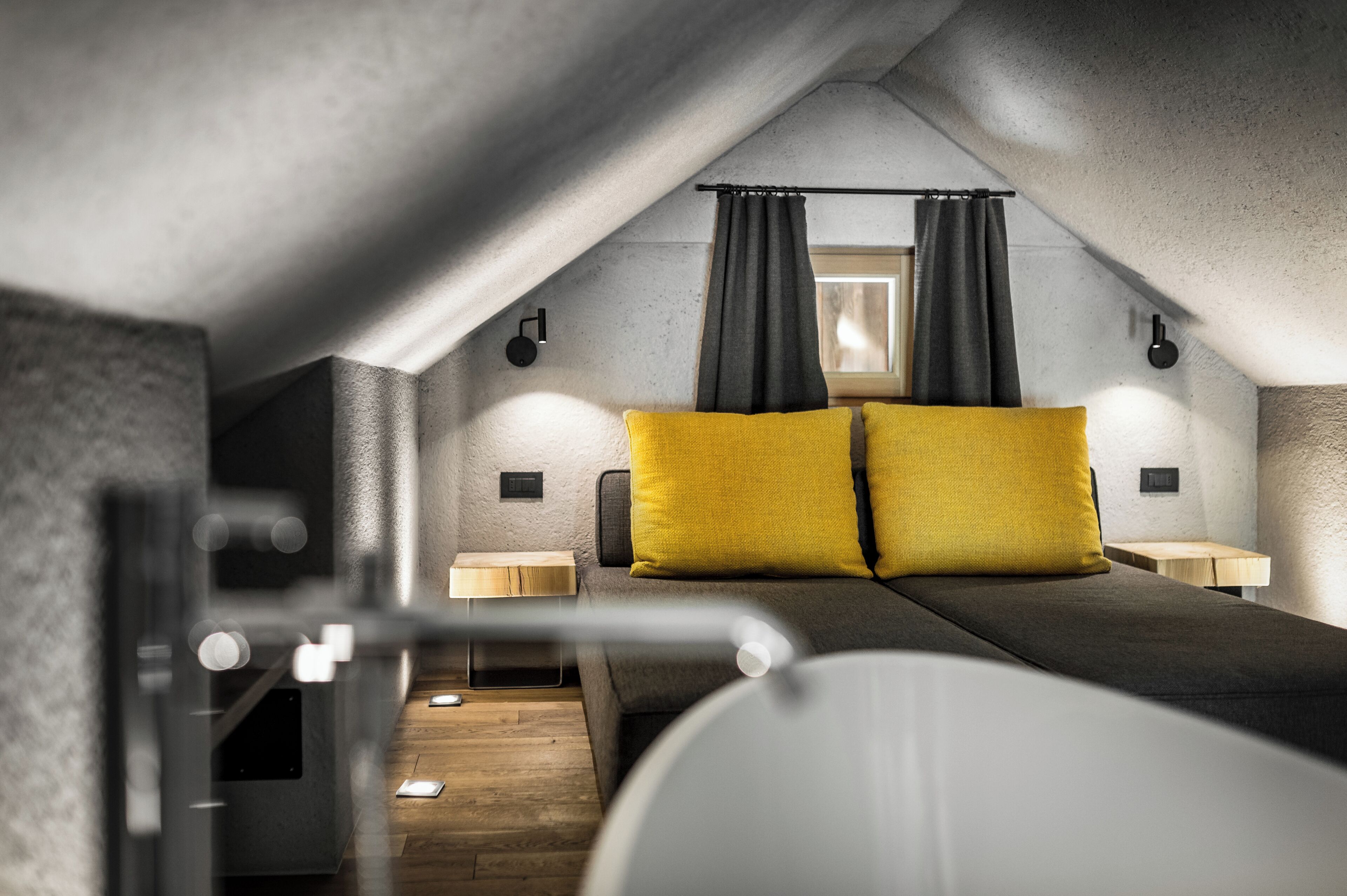 A modern, compact bedroom with a sloped ceiling, a bed with two yellow pillows, bedside lamps, and a window with curtains.