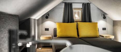 A modern, compact bedroom with a sloped ceiling, a bed with two yellow pillows, bedside lamps, and a window with curtains.