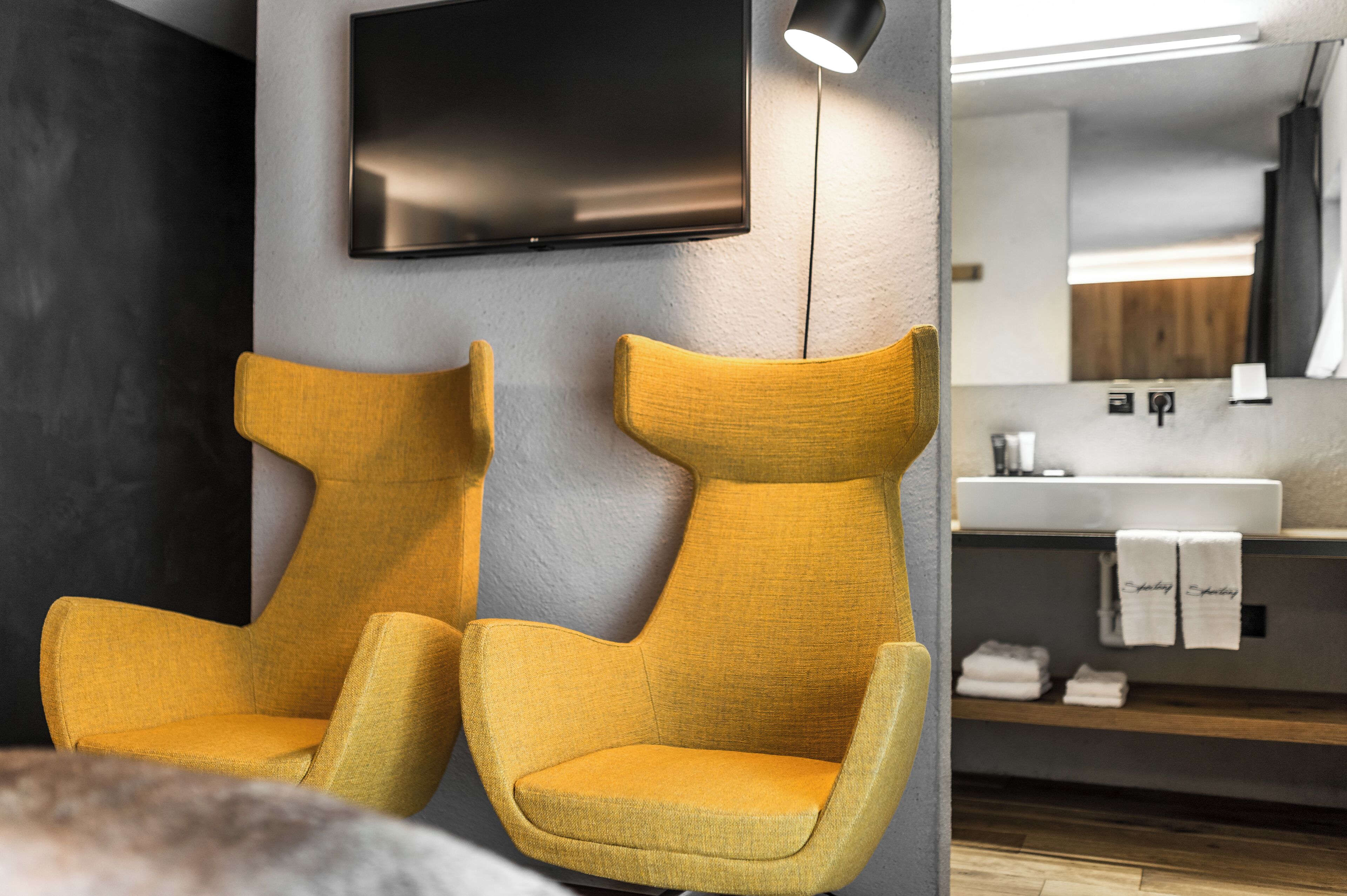A modern bathroom with two yellow chairs, a flat-screen TV mounted on the wall, and a vanity with a mirror and sink.