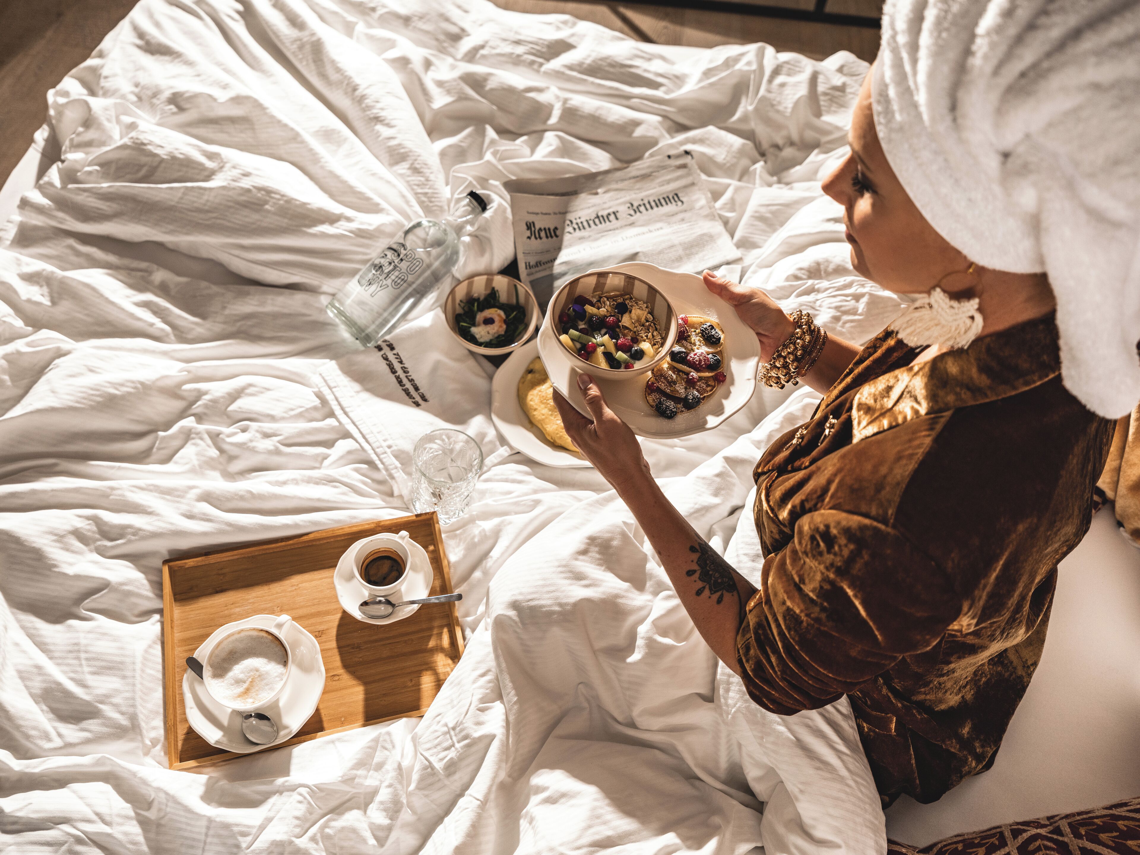Eine Person sitzt auf einem Bett mit einem Tablett, das eine Tasse Kaffee, eine Schüssel Obst und eine Zeitung enthält.