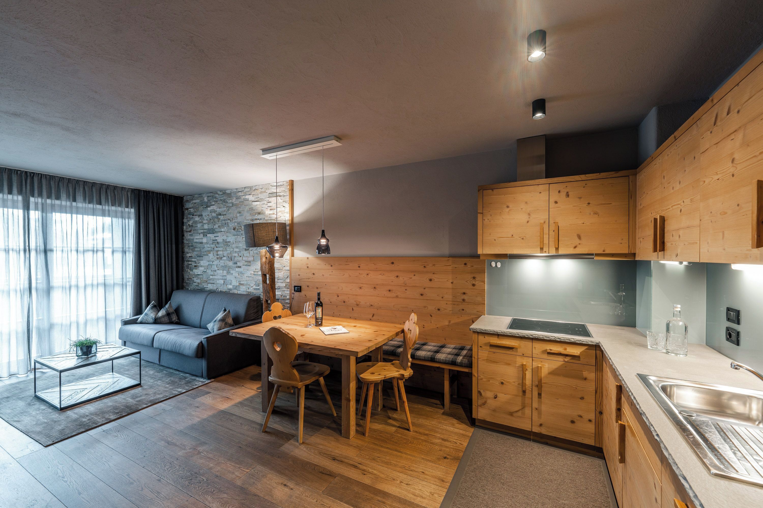 A modern living room with a grey sofa, a wooden dining table, and a kitchen area with wooden cabinets and a built-in oven.