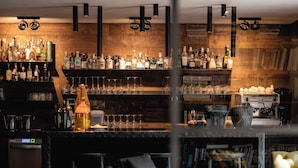 Zona de bar con pared de madera, estantes con botellas, vasos y una cafetera. Hay taburetes de bar y un área de asientos con una mesa y sillas.
