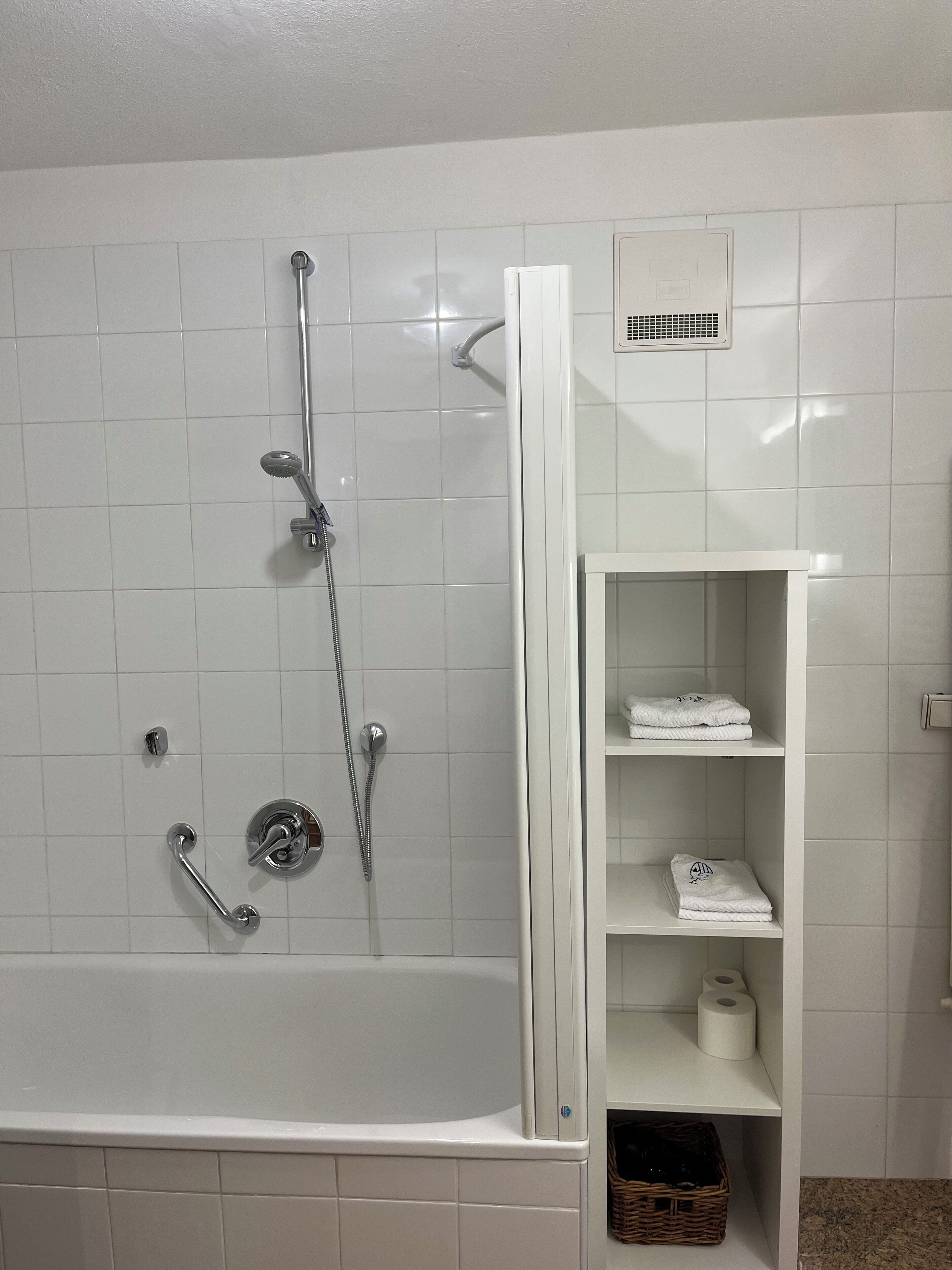 A modern bathroom with a bathtub, a shower with a handheld showerhead, and a white tiled wall. There is a white shelf with towels and a basket.