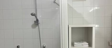 A modern bathroom with a bathtub, a shower with a handheld showerhead, and a white tiled wall. There is a white shelf with towels and a basket.
