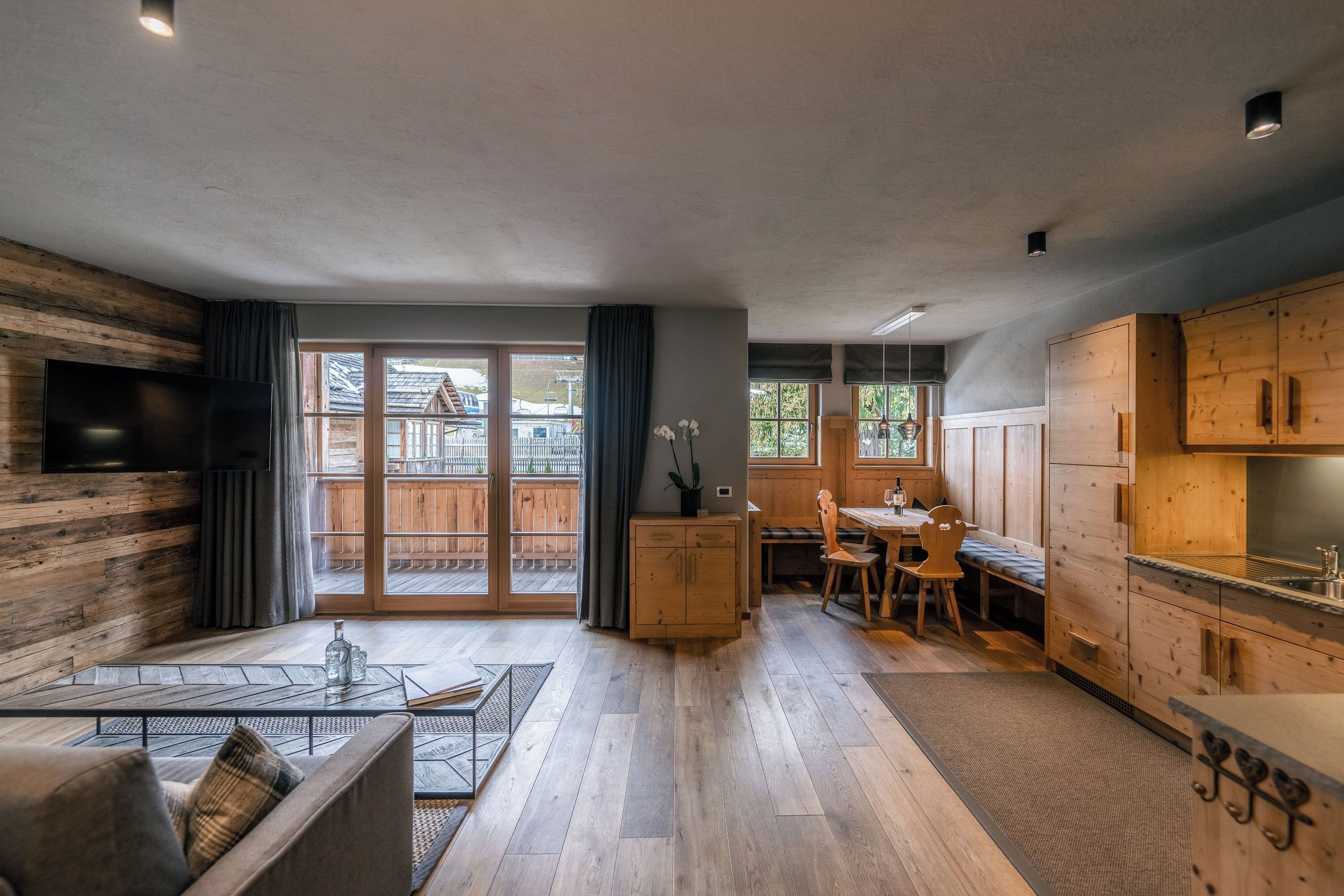 A modern interior with a wooden floor, a dining area, a kitchen with wooden cabinets, and a large window providing a view of the outside.