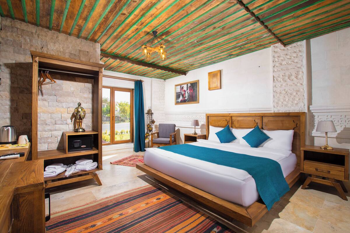Une chambre avec un grand lit, une tête de lit en bois, une table de chevet en bois, une commode, une armoire en bois, un luminaire suspendu et une vue sur l’extérieur.