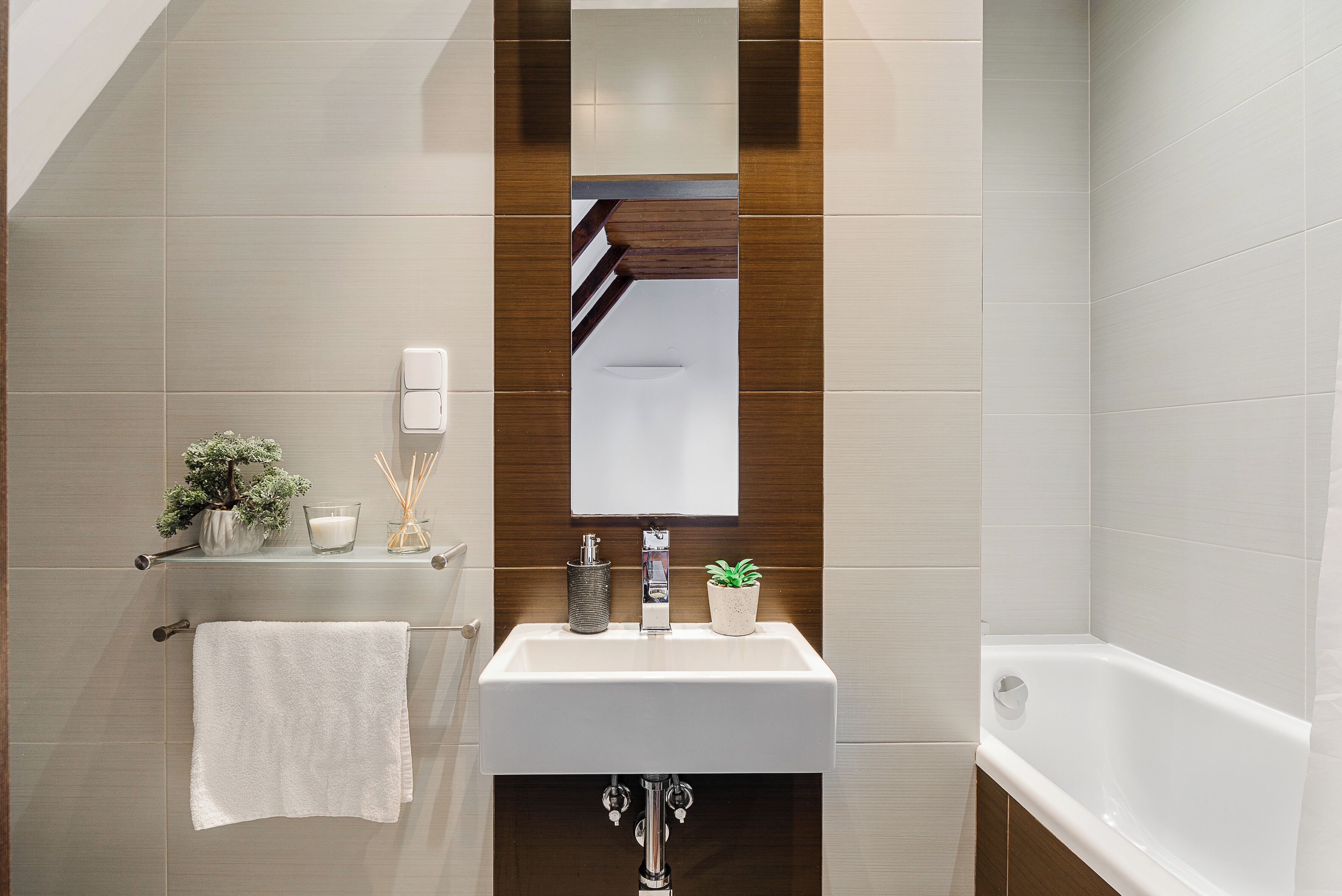 A modern bathroom with a white sink, mirror, and bathtub. There is a towel hanging on a rack, a small potted plant, and a candle on the sink.