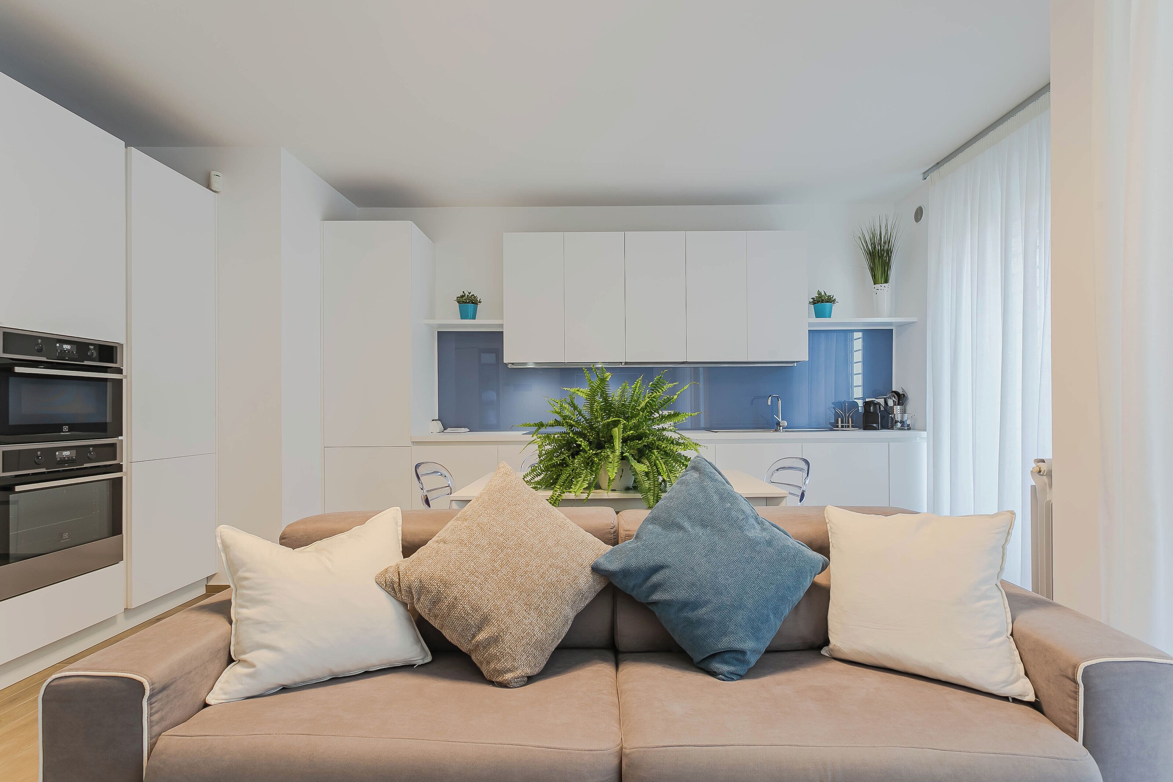 A modern living room with a beige sofa, three cushions, a kitchen in the background, and a potted plant on the counter.