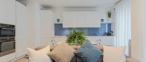 A modern living room with a beige sofa, three cushions, a kitchen in the background, and a potted plant on the counter.