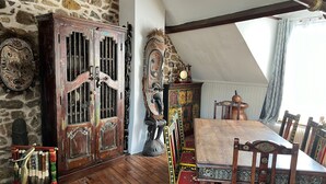 Um quarto com parede de pedra, um armário de madeira com detalhes entalhados, uma cadeira de palha tradicional, uma cama de madeira com cabeceira estampada e uma mesa de madeira com cadeiras.
