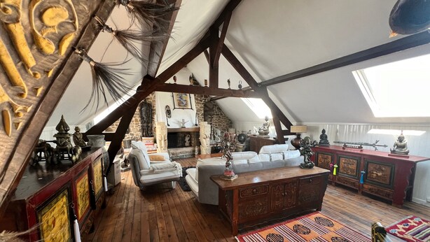 Quarto estilo loft com vigas de madeira aparentes, teto alto e claraboia. Há um sofá branco, um armário de madeira e vários objetos decorativos, incluindo estátuas e uma figura de Buda.