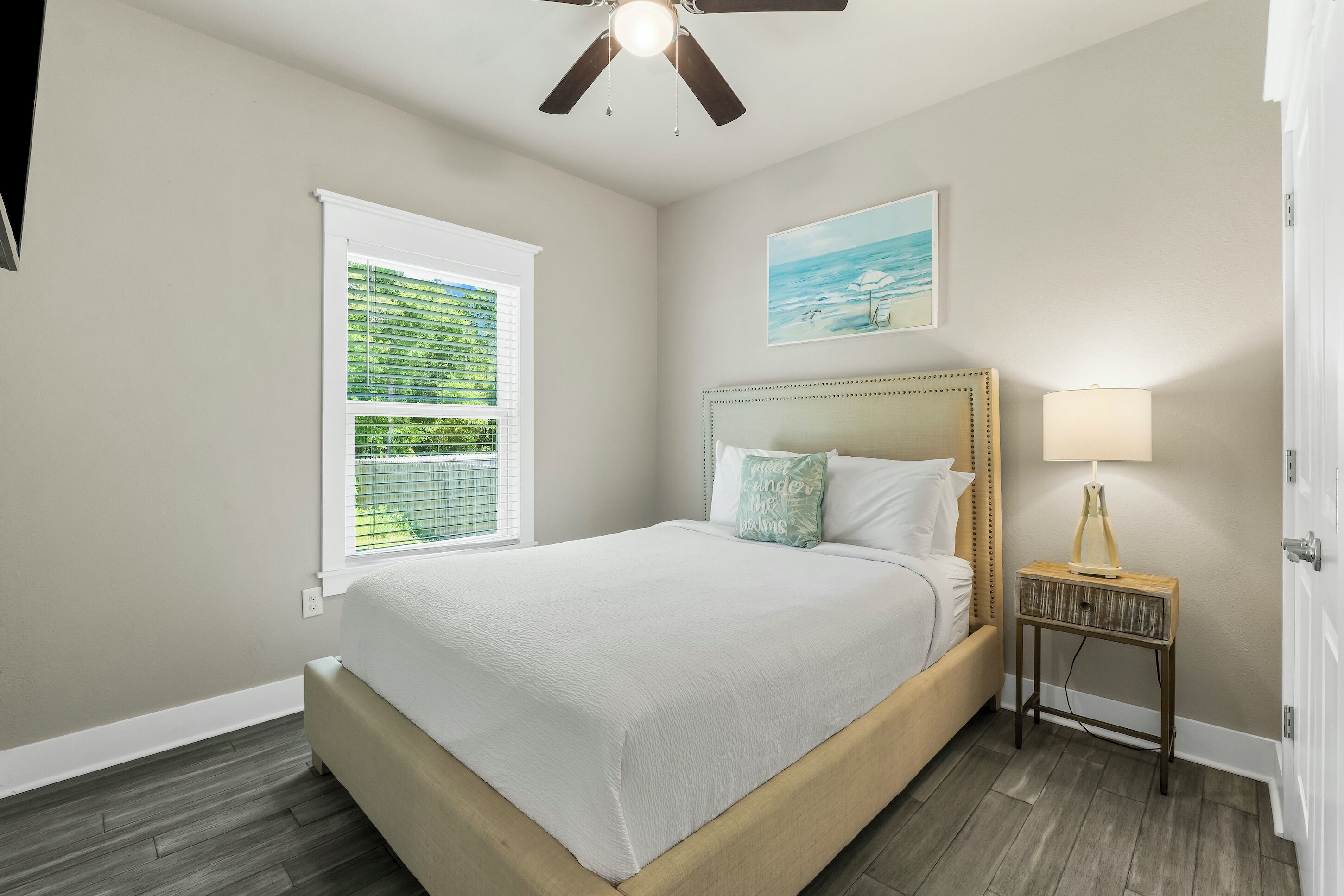 A bedroom with a bed, a window with blinds, a ceiling fan, a bedside table with a lamp, and a painting on the wall.